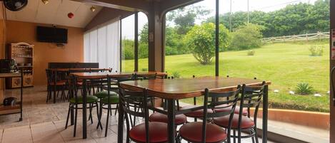 A dining area with a large table and chairs, a television, and a view of a grassy landscape through large windows.