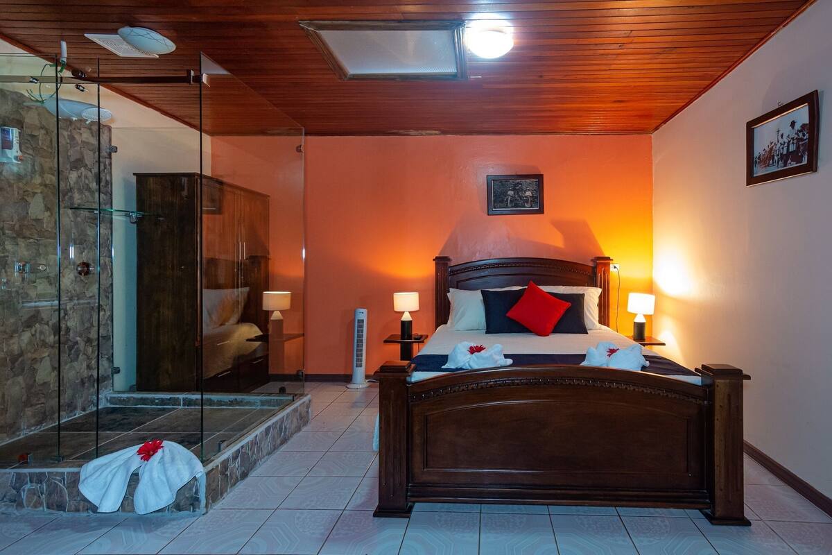 A hotel room with a wooden ceiling, a bed with a red pillow, a nightstand with a lamp, and a picture on the wall.