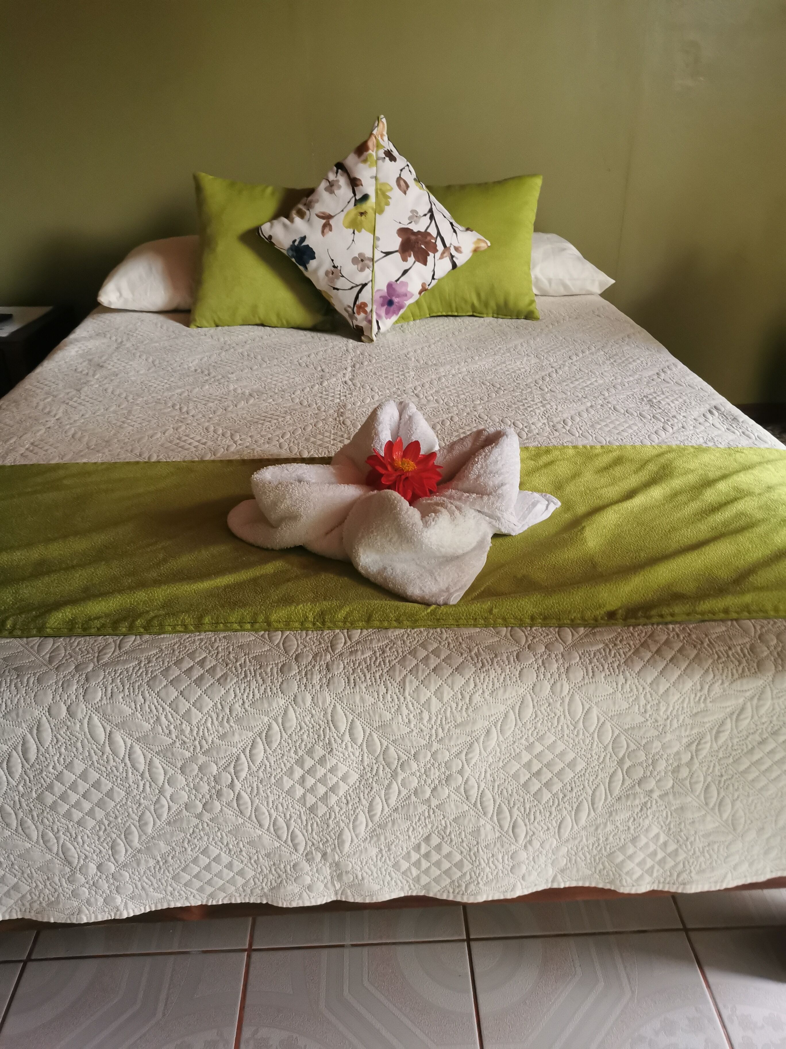Ein ordentlich bezogenes Bett mit einem floralen Kissen, einer grünen Tagesdecke und einem Handtuch mit Blumenmuster.