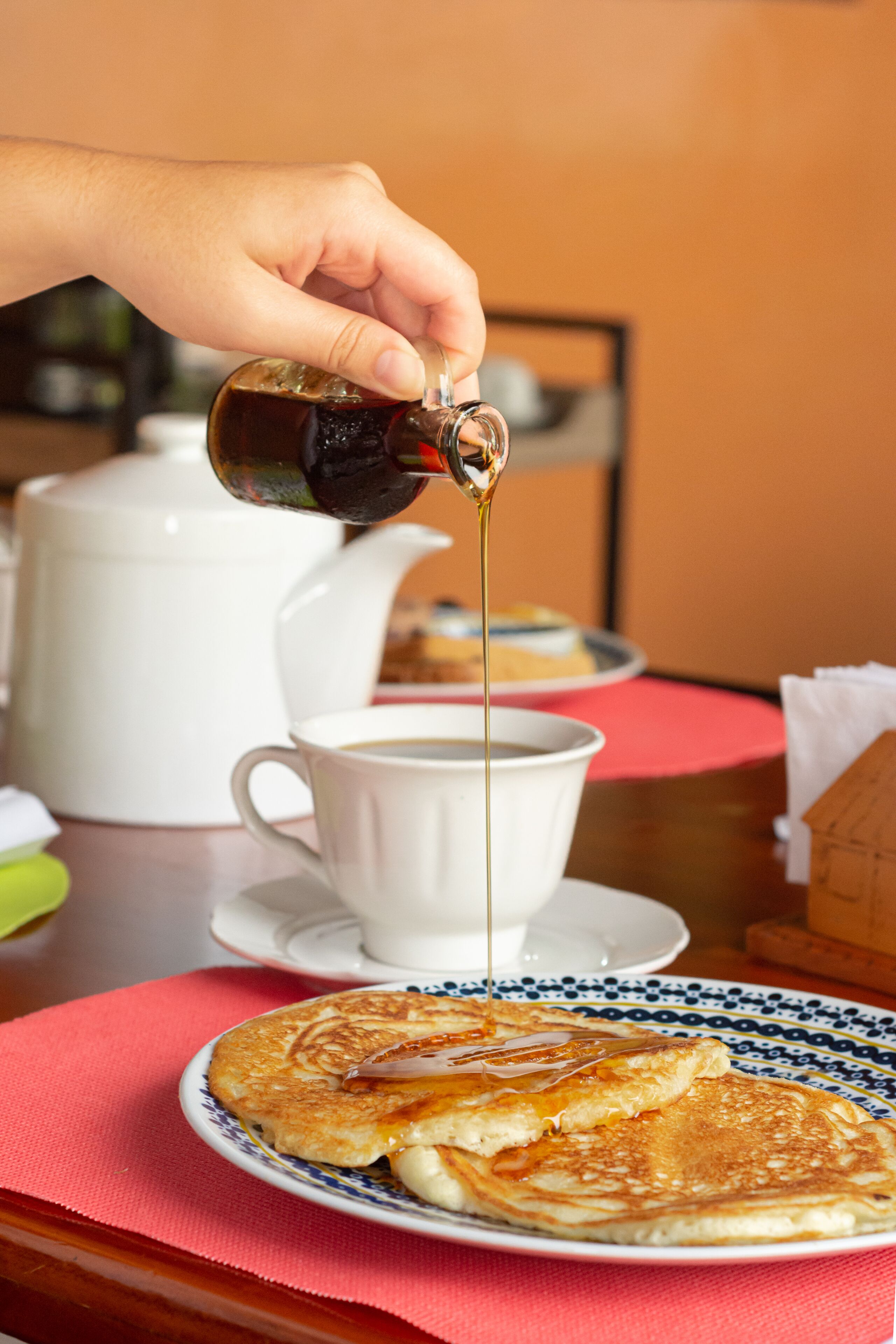 A hand pouring syrup onto pancakes on a plate, with a cup of coffee and a teapot in the background.