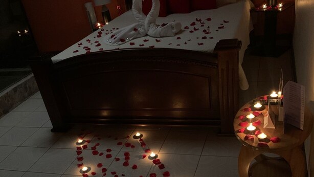 A dimly lit bedroom with a bed adorned with rose petals and candles, a wooden nightstand with a lamp, and a tiled floor. - Nafre House (Monteverde)