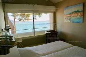 A bedroom with a large window offering a view of the sea and a painting of a coastal scene.