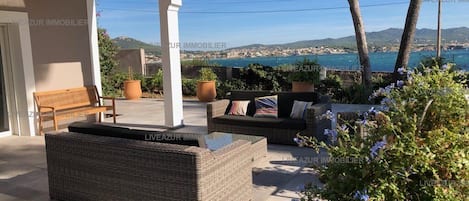 Un espacio de asientos al aire libre con mobiliario de mimbre y plantas en macetas, con vistas a un paisaje costero.