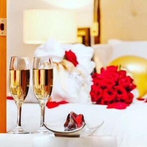 A neatly arranged hotel room with a bed, two champagne flutes, a plate with a dessert, and a bouquet of red roses.