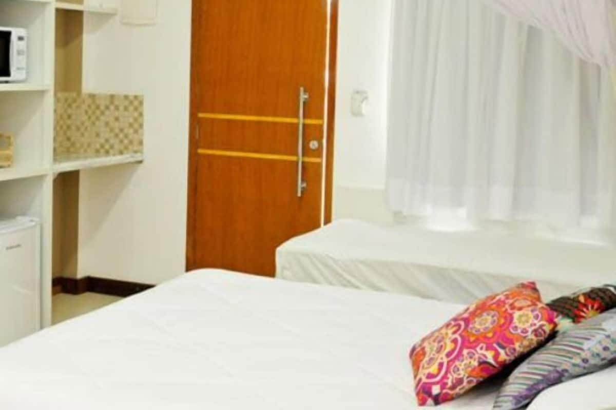 Quarto de hotel com porta de madeira, cama com roupa de cama branca, mesa de cabeceira, prateleira com micro-ondas e janela com cortinas finas.