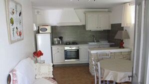 Une cuisine compacte avec des meubles blancs, un réfrigérateur, un four à micro-ondes et une cuisinière. On y trouve une table à manger avec des chaises et un petit îlot central. Un canapé blanc agrémenté de coussins à motifs floraux est placé contre le mur.