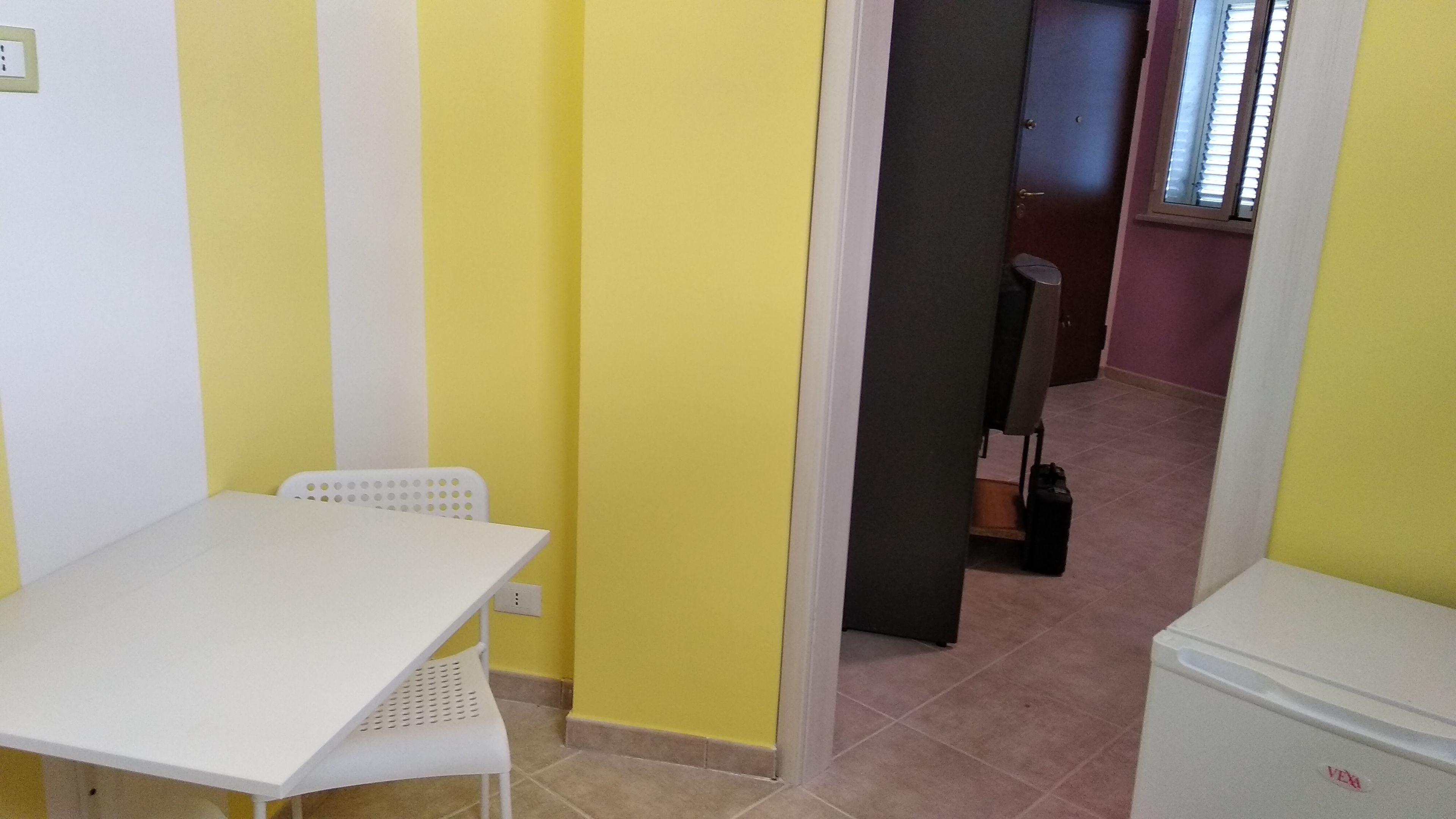 Una habitación pequeña con una mesa y una silla blancos, paredes amarillas, una puerta que da a otra habitación y un refrigerador pequeño.