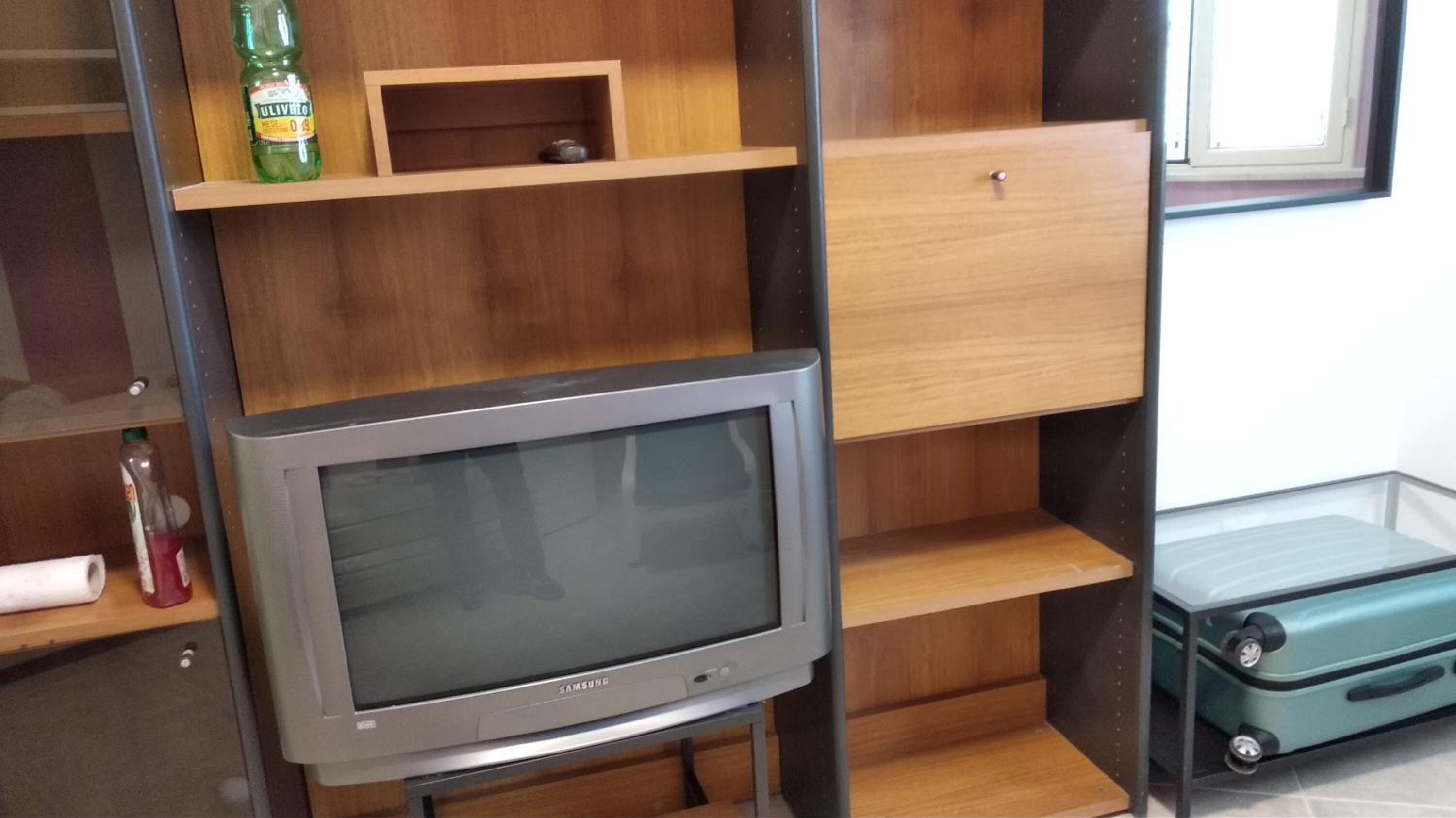 A room with a Samsung TV on a stand, wooden shelves, a small cabinet, and a suitcase.