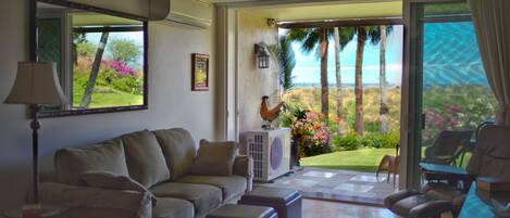 A living room with a sofa, ottomans, a chair, a lamp, and a view of a garden with palm trees and flowers.
