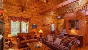 A cozy log cabin interior with a fireplace, leather sofa, wooden dining table, and a staircase leading to an upper level. - Baird's Creek Cabin  Cozy Cabin, Close to Golf, Creekside (Vilas)