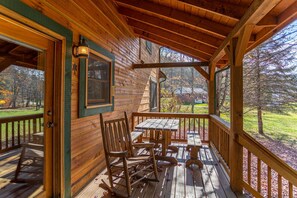 Relax on the porch with a wooden table and chairs, and a comfortable rocking chair, surrounded by a serene grassy area with trees.