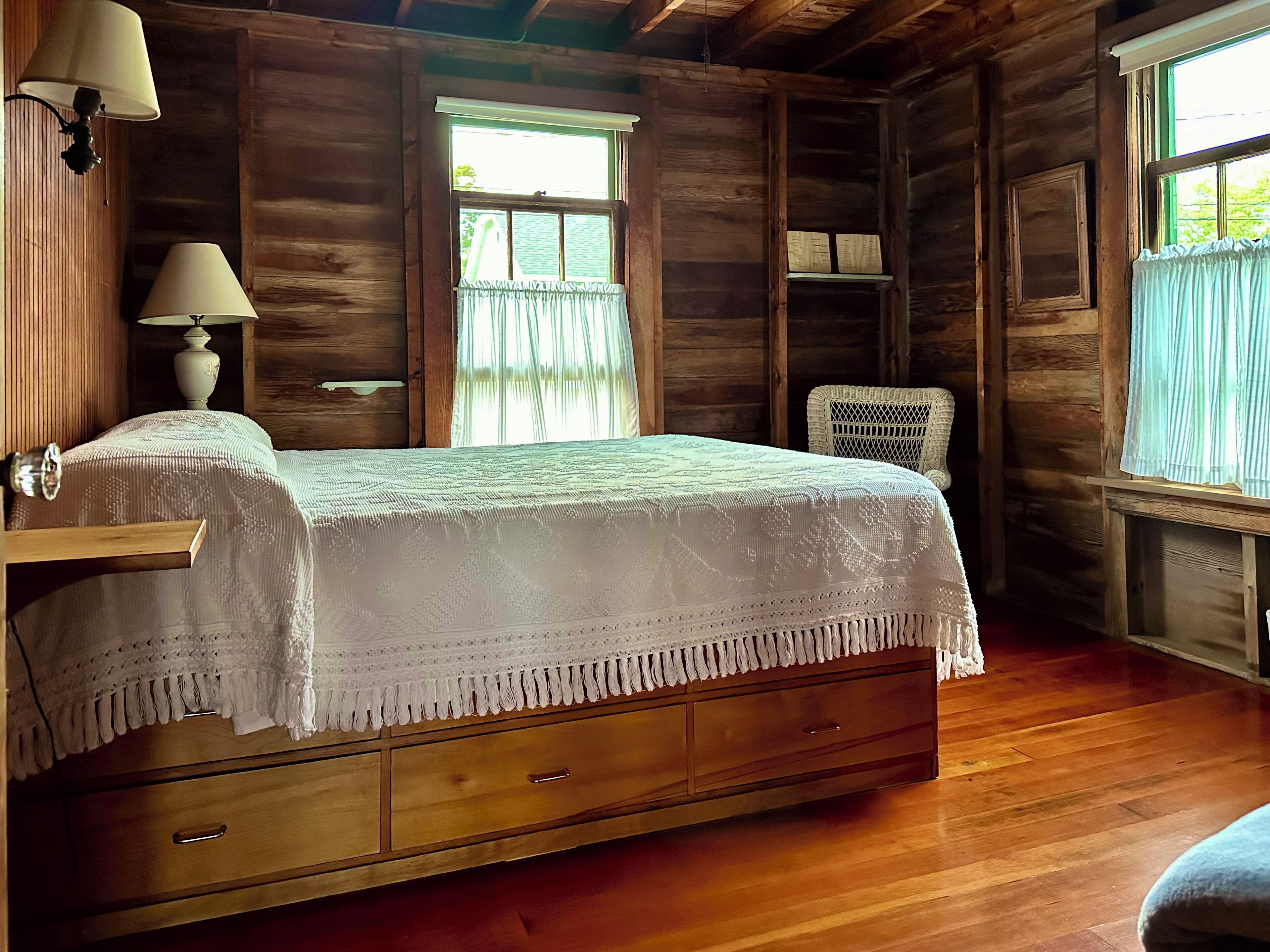 Quarto de cabana de madeira com cama, duas luminárias de cabeceira, uma cadeira de vime e uma janela com cortinas.