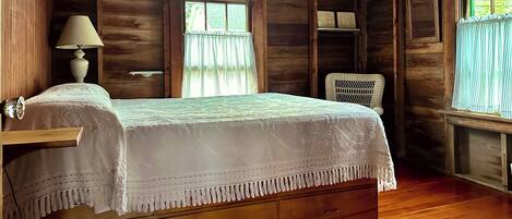 Quarto de cabana de madeira com cama, duas luminárias de cabeceira, uma cadeira de vime e uma janela com cortinas.