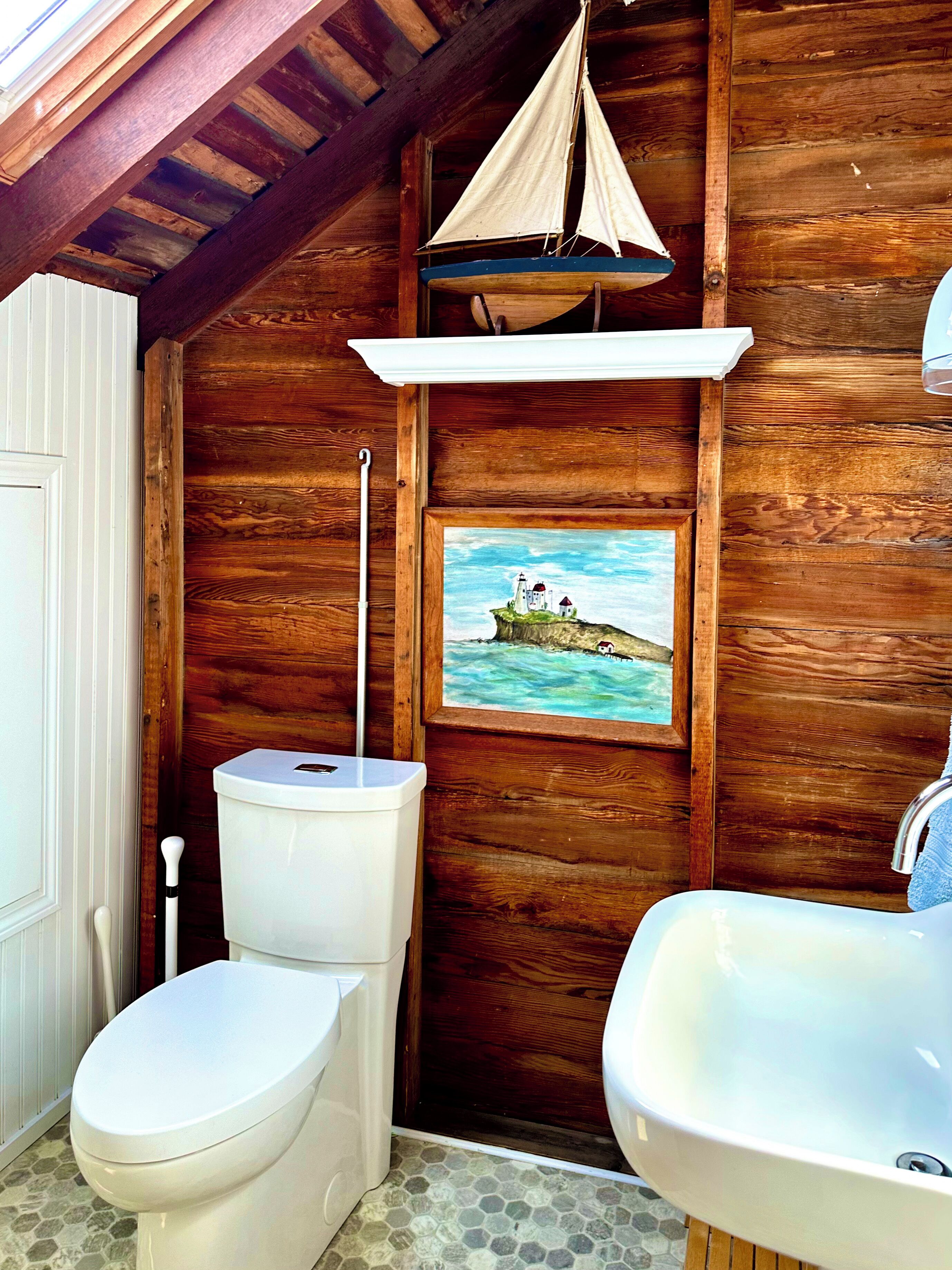 Ein Badezimmer aus Holz mit Toilette, Waschbecken und einem Gemälde eines Leuchtturms an der Wand.