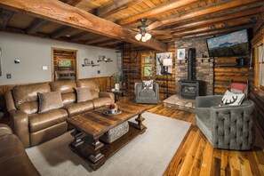 Ein Interieur einer Blockhütte mit einem Ledersofa, einem grauen Sessel, einem Holztisch, einem Kamin und einem an der Wand befestigten Fernseher.