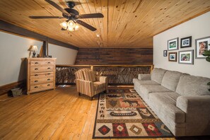 A cozy room with a wooden ceiling, a ceiling fan, a sofa, a chair, a wooden cabinet, and a rug. - Riverside Retreat | Private Riverfront, Hot Tub, Game Room & Fire Pit (Boone)