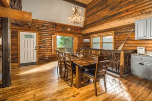A wooden cabin interior with a dining table, chairs, a chandelier, and a view through the window. - Riverside Retreat | Private Riverfront, Hot Tub, Game Room & Fire Pit (Boone)