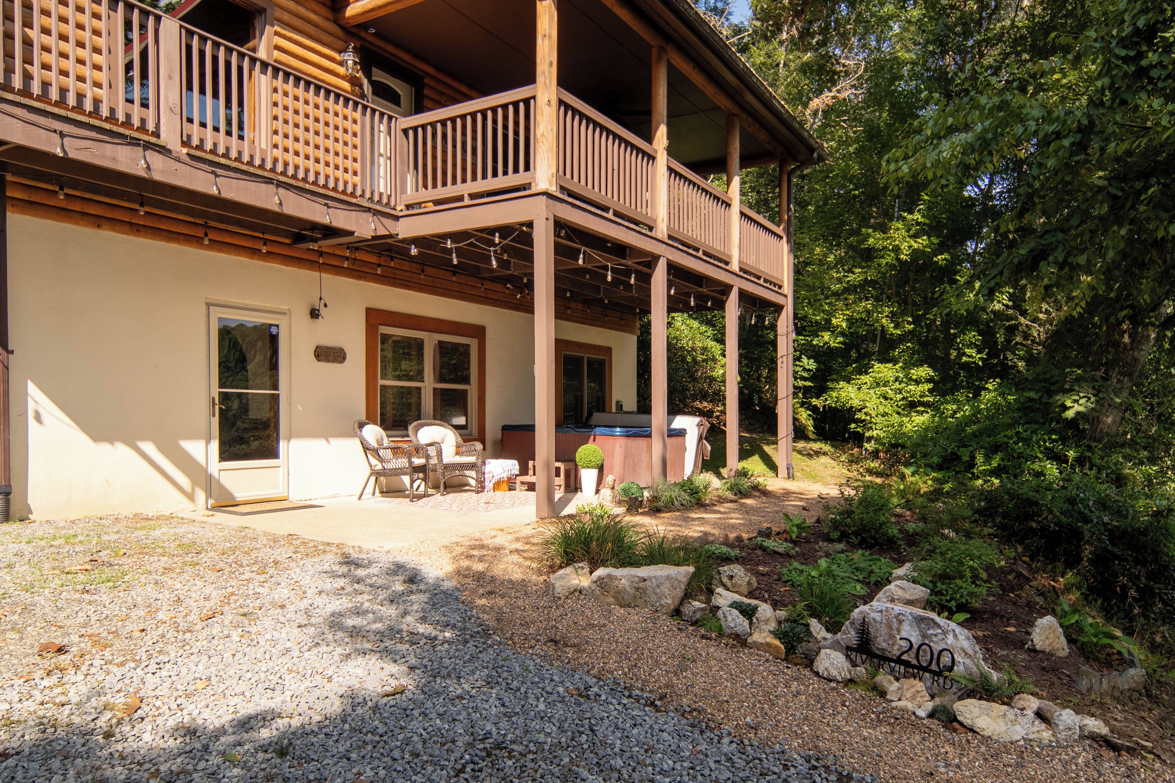 A two-story wooden cabin with a balcony, a covered patio with outdoor furniture, and a gravel driveway.