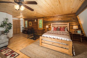 A log cabin bedroom with a wooden bed, a ceiling fan, a window with blinds, and a plant. - Riverside Retreat | Private Riverfront, Hot Tub, Game Room & Fire Pit (Boone)