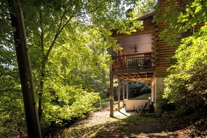 A wooden cabin with a balcony, surrounded by dense green foliage. - Riverside Retreat | Private Riverfront, Hot Tub, Game Room & Fire Pit (Boone)
