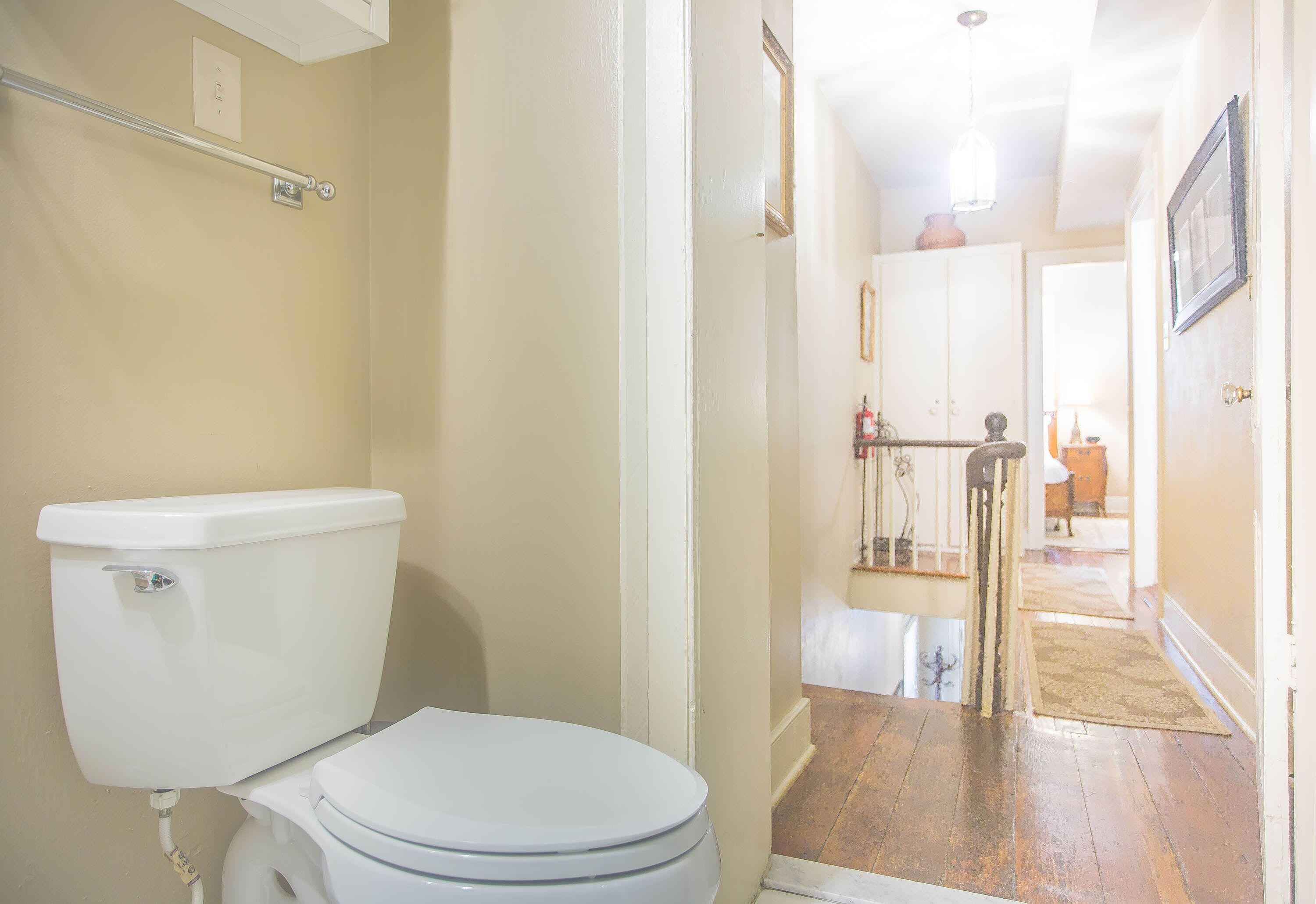 A bathroom with a white toilet, a handrail, and a door leading to a hallway with a wooden floor and a rug.