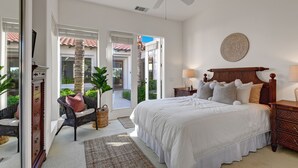 Une chambre avec un grand lit, un ventilateur de plafond, une table de chevet, un fauteuil en osier et une vue sur l’extérieur.