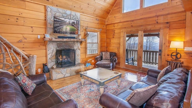 A cozy log cabin interior with a stone fireplace, leather sofas, and a large window overlooking a deck. - Large Cabin Sitting at 4,000 Feet Elevation (Zionville)