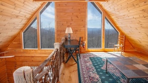 A cozy cabin with a wooden interior, a dining table, a chair, a lamp, and a view of the mountains through large windows. - Large Cabin Sitting at 4,000 Feet Elevation (Zionville)