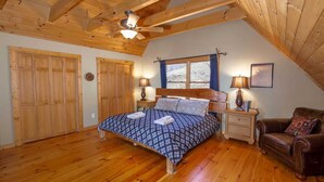 A bedroom with a wooden ceiling, a bed with a patterned bedspread, two bedside lamps, a wooden nightstand, a brown leather armchair, and a window with curtains. - Large Cabin Sitting at 4,000 Feet Elevation (Zionville)
