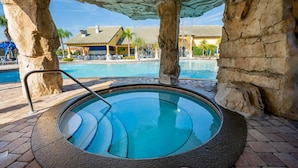 A hot tub with a stone surround, a pool in the background, and a building with a tiled roof. - Beautiful Town Home in Gated Community with Extensive Facilities (Kissimmee)