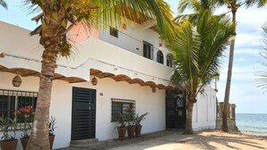 Un edificio de dos pisos con fachada blanca, contraventanas negras y el número '66' en la pared. Hay plantas en macetas y una palmera frente al edificio.