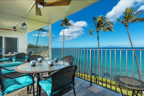 A covered outdoor area with a table and chairs, a view of the ocean, and palm trees.