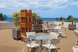 An outdoor seating area with tables and chairs, a wooden decorative panel, and a view of the ocean.