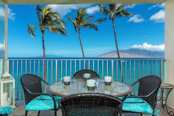 A balcony with a round glass table, four wicker chairs with blue cushions, and a view of the ocean and mountains.