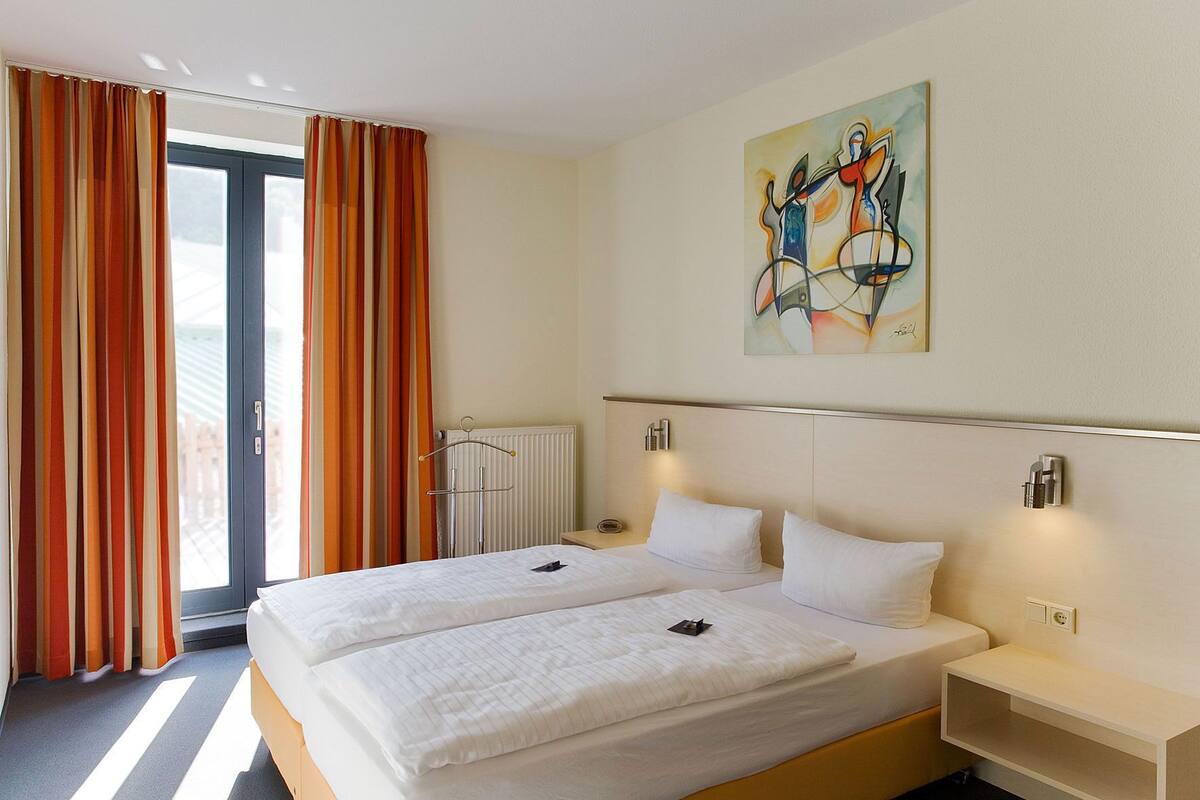 A hotel room with a large bed, a radiator, a painting on the wall, and a window with curtains.