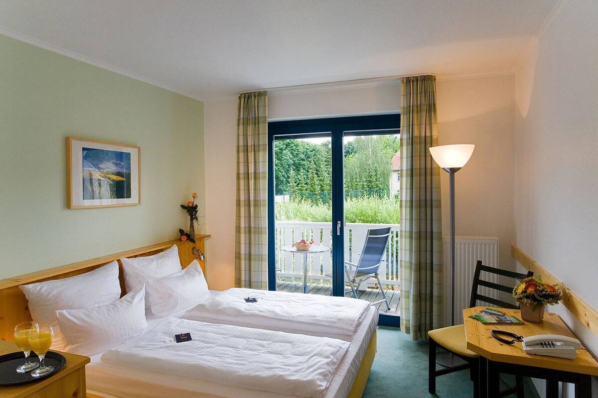 A hotel room with two beds, a balcony with a table and chairs, a framed picture on the wall, and a lamp on the table.