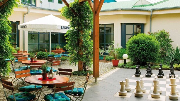 An outdoor patio area with round tables and chairs, a chessboard, and greenery. - aktiv Sporthotel (Pirna)