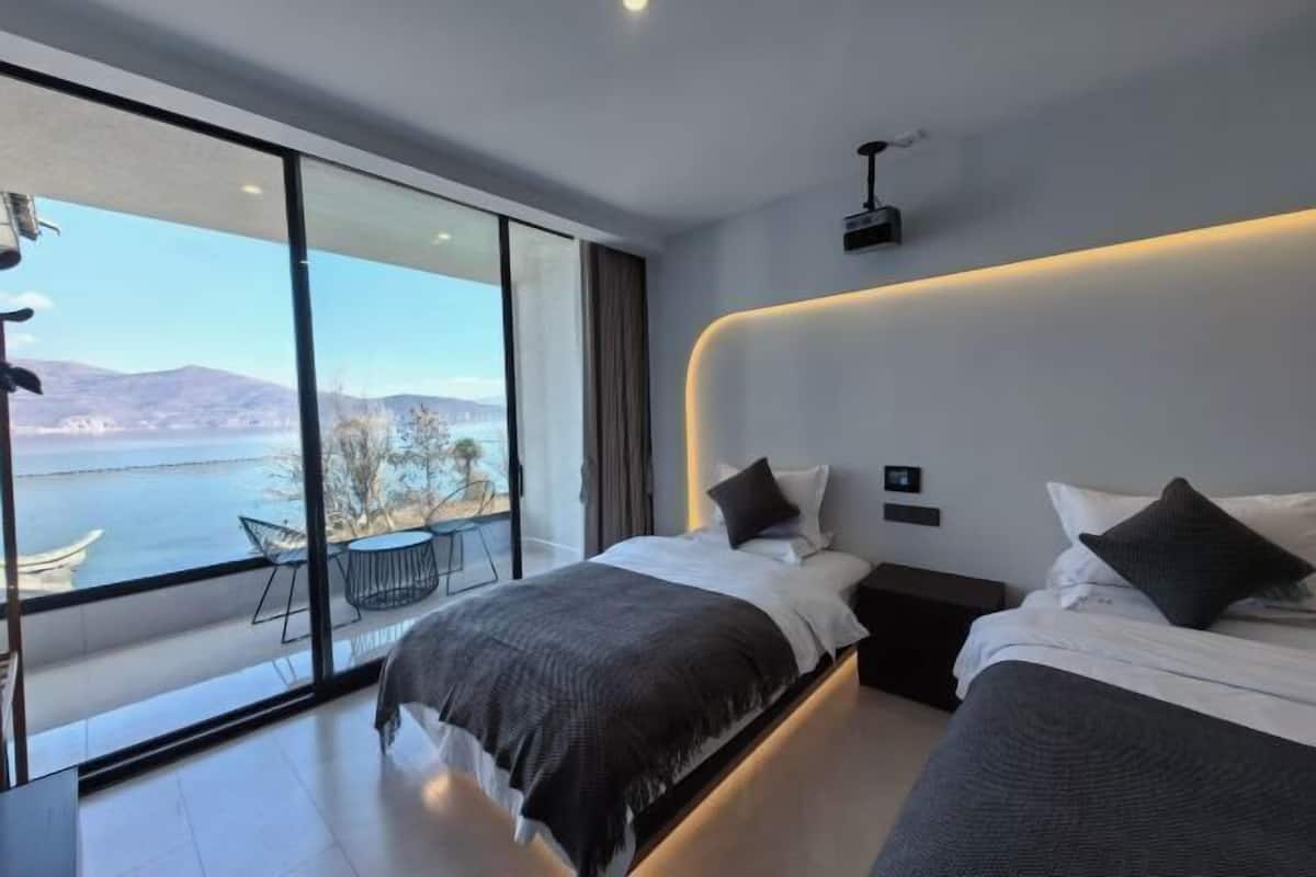 A modern bedroom with two beds, a large window with a view of the sea, and a small table with chairs outside.