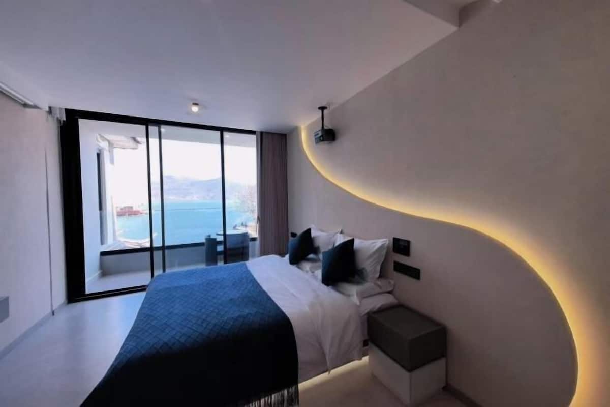 A modern bedroom with a large bed, a bedside table, and a view of the sea through a sliding glass door.