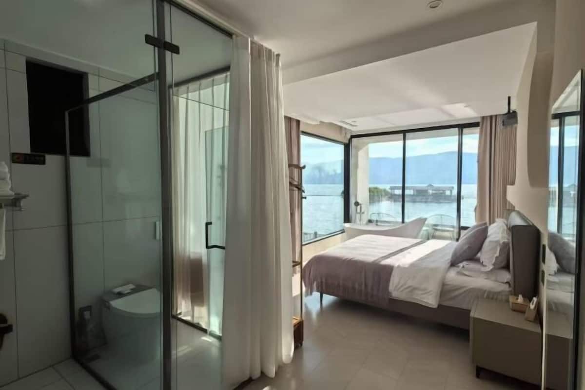 A modern hotel room with a large bed, a glass shower enclosure, and a balcony with a view of the water.