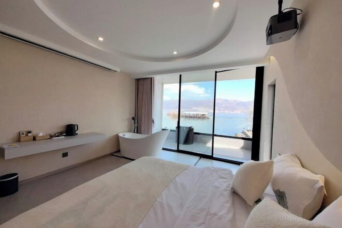 A modern bedroom with a large bed, a built-in bathtub, and a balcony with a view of the sea.
