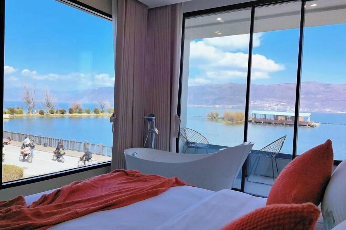 A modern hotel room with a large window, a bathtub, and a bed with pillows.
