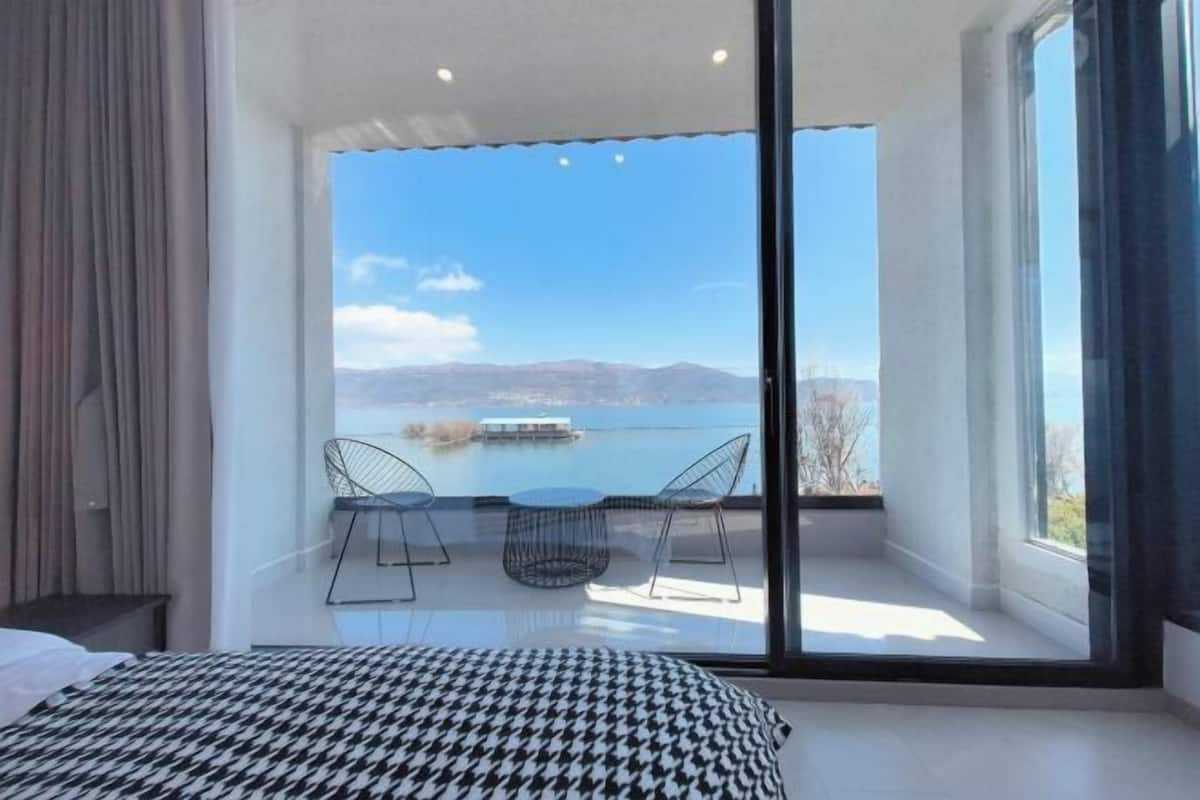 A bedroom with a large sliding glass door leading to a balcony with a view of a lake and mountains.