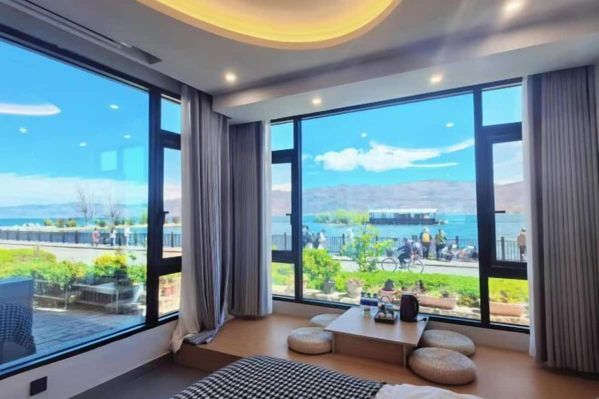 A modern hotel room with large windows offering a view of a coastal area, a small seating area, and a bed.
