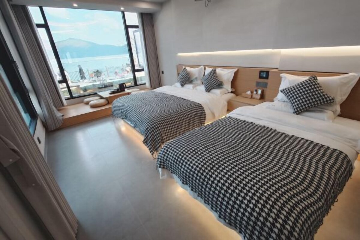 A hotel room with two beds, a large window with a view, and a bedside table with a lamp.
