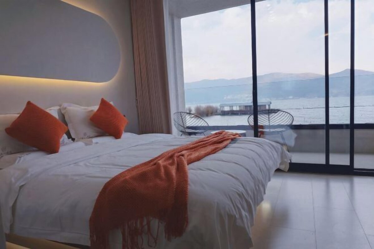 A modern bedroom with a large bed, orange pillows, and a view of a lake and mountains.