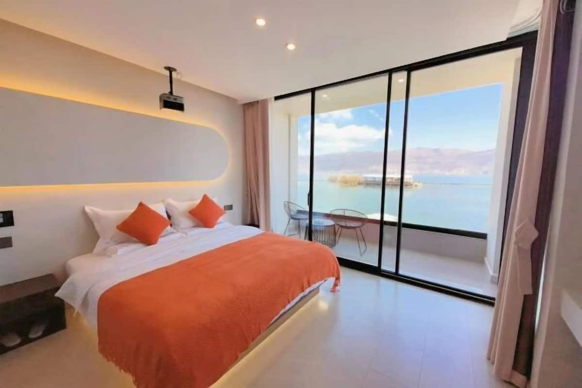 A modern bedroom with a large bed, a view of a body of water through floor-to-ceiling windows, and a balcony with a table and chairs.