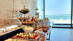 Un bufé con diversos alimentos, entre ellos frutas, verduras y pasteles, en una habitación con un ventanal que ofrece vista al mar.