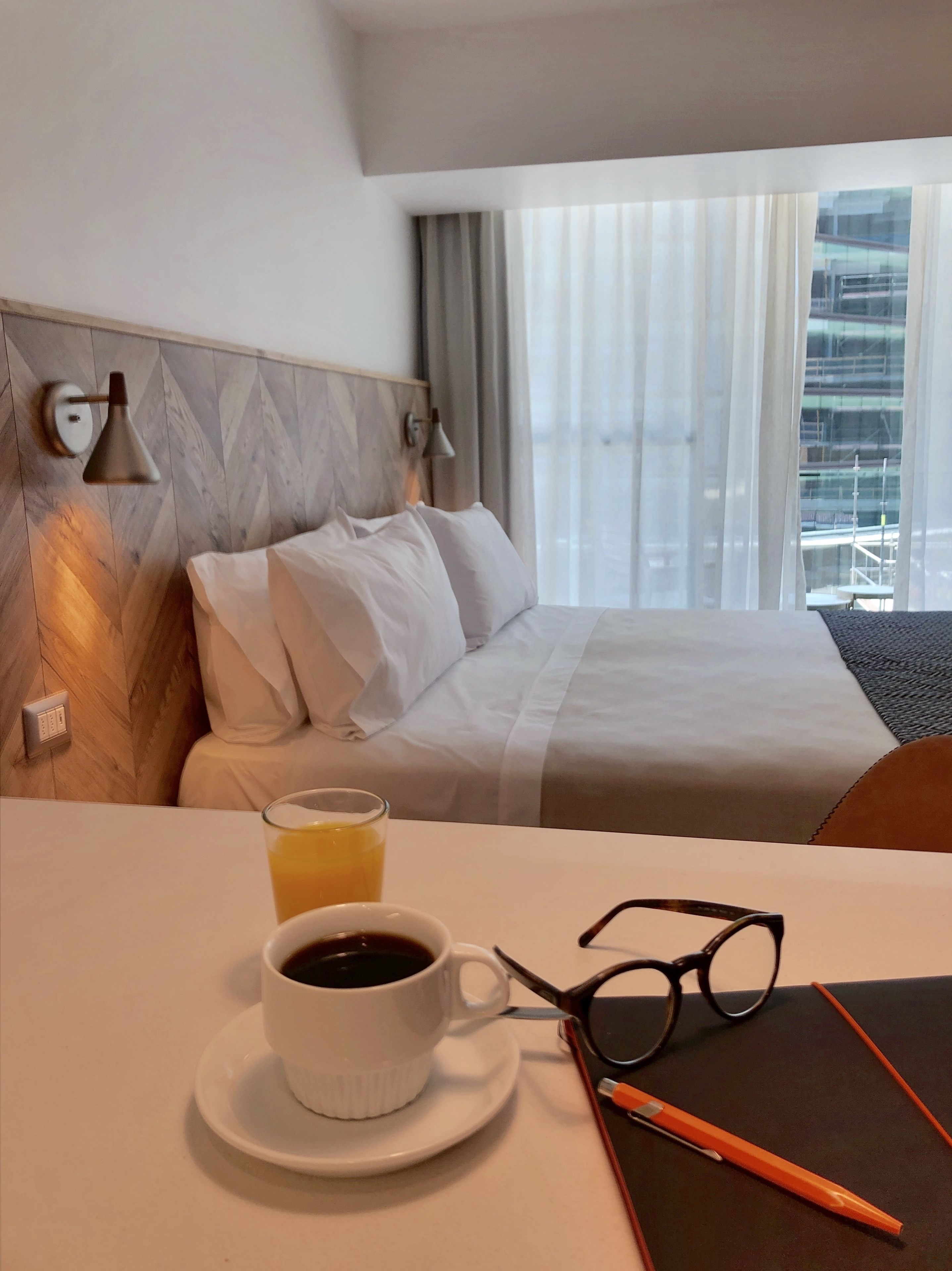 Et hotelværelse med en seng, et natbord med en kop kaffe og glas, en lampe og et vindue med gardiner.