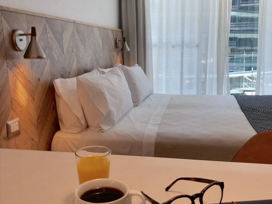 Et hotelværelse med en seng, et natbord med en kop kaffe og glas, en lampe og et vindue med gardiner.